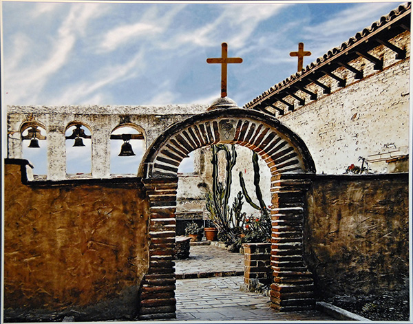 Looking through arched entrance to mission courtyard with bells in background and cross on top of curved archway entrance
